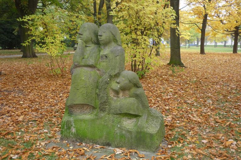 auf der Wiese steht eine Sandsteinskulptur mit drei teils nur angedeuteten Personen